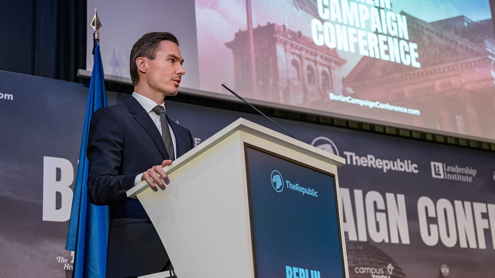a man in a suit speaking at Berlin Campaign Conference (presentation in the background)